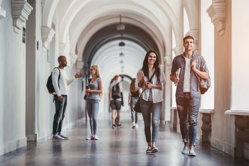 Happy multiracial teenagers walk on university hall background, happy male and female students walking and communicating, having coffee break in university hall.